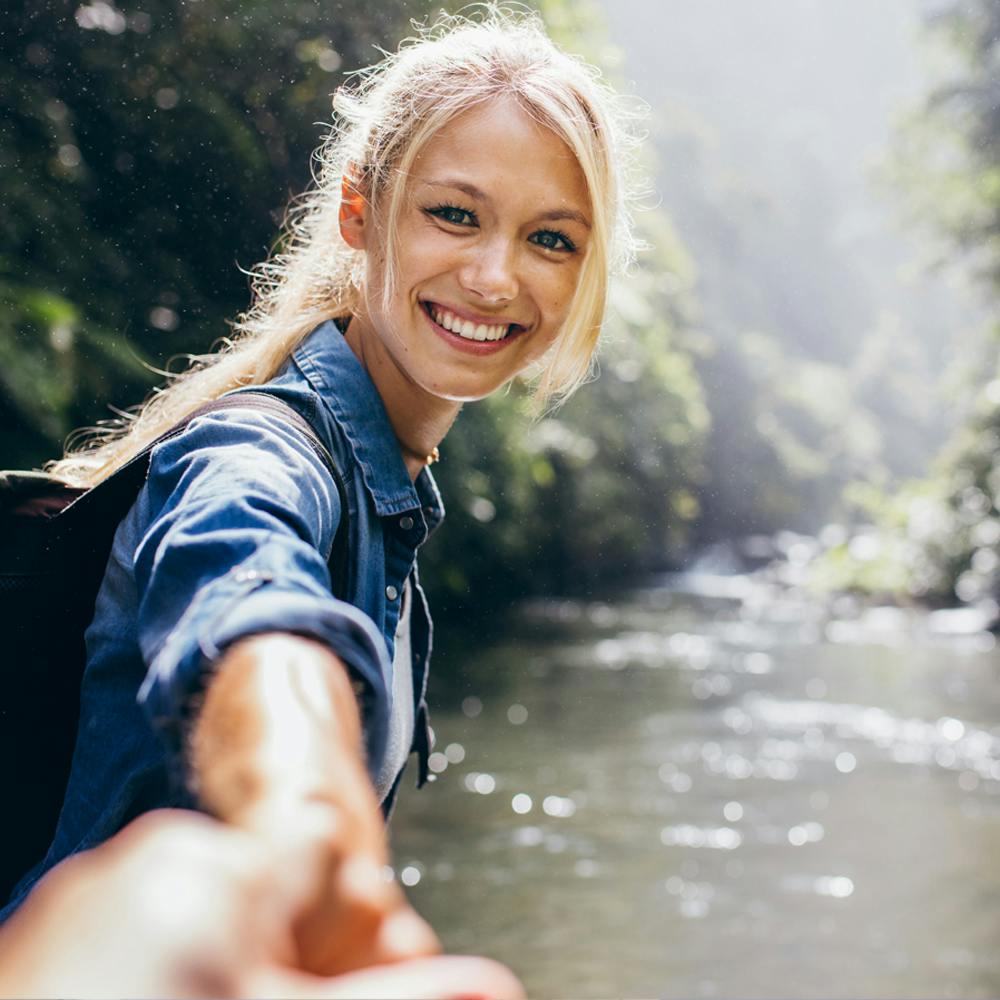 Eine junge Frau mit blonden Haaren und Rucksack, die in die Kamera lächelt und eine Hand ausstreckt, mit einem Fluss und Bäumen im Hintergrund.