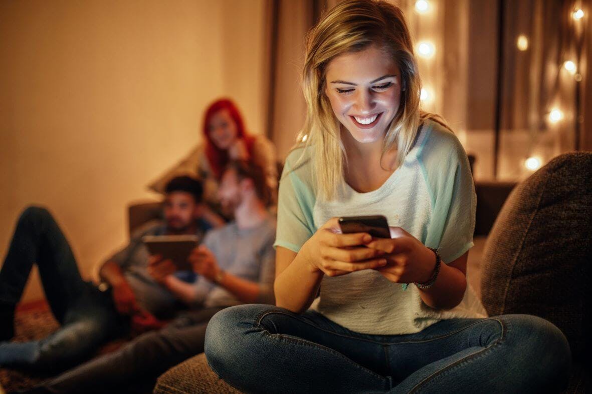 Eine junge Frau mit blonden Haaren sitzt im Schneidersitz auf einem braunen Sofa und schaut lächelnd auf ihr Smartphone. Das helle Licht des Bildschirms beleuchtet ihr Gesicht. Im Hintergrund, auf demselben Sofa, liegen und sitzen zwei weitere Personen, ein Mann mit Bart, der auf ein Tablet schaut, und eine Frau mit roten Haaren, die in Richtung der Frau mit dem Smartphone blickt. Die Szene ist warm beleuchtet, wahrscheinlich durch eine Lampe im Hintergrund, und wirkt gemütlich.